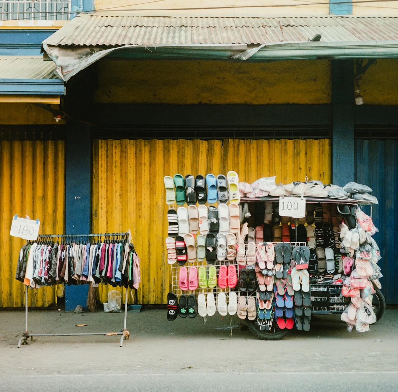 A vibrant street market stall showcasing slippers and clothes against a corrugated wall backdrop.