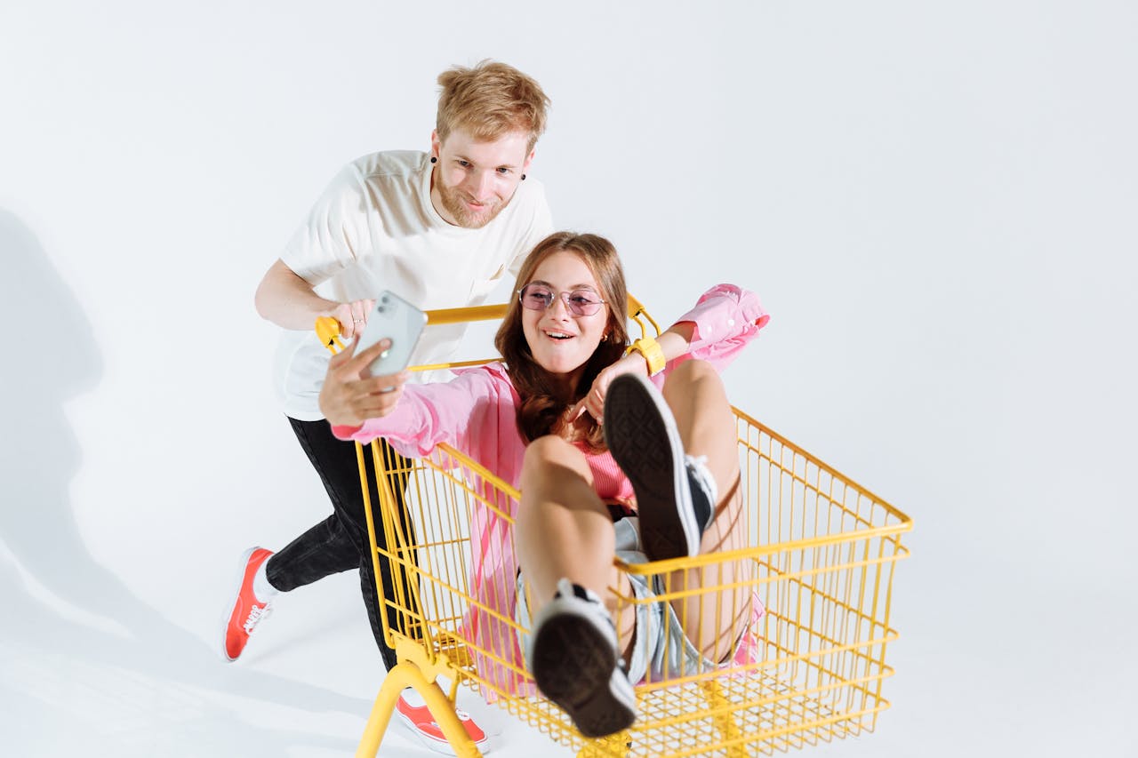 Young woman enjoys a fun shopping cart ride, taking a selfie with a young man pushing.